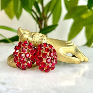 May include: A pair of red rhinestone earrings in a heart shape. The earrings are on a gold-colored surface.