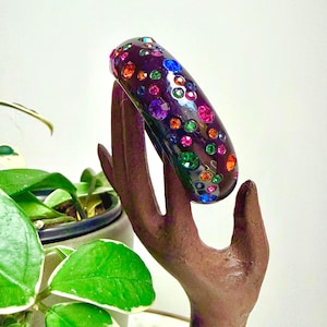 May include: A dark purple, smooth, rounded bangle bracelet featuring an array of embedded, multi-colored, circular faceted crystals in shades of blue, green, orange, and pink, arranged in a scattered, non-uniform pattern across the surface.