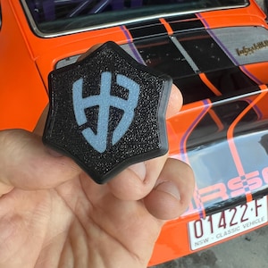 May include: A black five-pointed star-shaped key fob with a white stylised letter 'H' logo. The key fob is being held in a hand in front of a bright orange, black, and purple car.