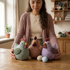 Könnte beinhalten: Drei handgestrickte Hühnerfiguren in den Farben Salbeigrün, Altrosa und Lavendel. Jedes Huhn hat einen kontrastierenden dunkelroten Kamm und Kehllappen, einen gelben Schnabel und kleine dunkle Augen. Zwei kleine, runde, gestrickte Eier liegen davor.