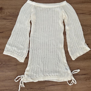 May include: Off-white, open-knit tunic with long, flared sleeves. The tunic has a boat neck and ties at the sides. The lightweight, textured fabric suggests a beach cover-up or summer garment.