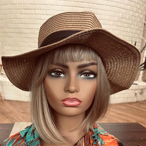 May include: A wide-brimmed, woven straw hat with a black band. The hat is a light brown color and has a slightly curved brim. The hat is on a mannequin head with blonde hair and a colorful shirt.