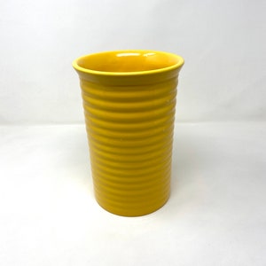 May include: A yellow ceramic tumbler with a ribbed design. The tumbler has a cylindrical shape and a slightly flared rim. The exterior is a solid, bright yellow colour. The tumbler is approximately 15 cm tall.