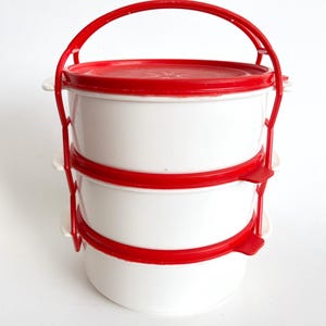 May include: A three-tiered white and red food carrier with a red handle. Each tier is a round container with a red lid and a red band around the middle. The carrier is designed for easy transport.