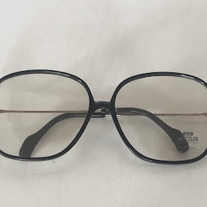 May include: Black-framed eyeglasses with a rounded square shape. The frames have a gold-colored bridge and temples. The lenses are clear. The glasses have a label with the text "MATZLER" on the right lens.