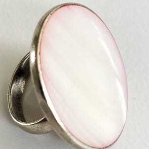 May include: Oval-shaped ring with a large, white and pink stone set in a silver-toned metal band. The stone has a pearlescent sheen, and the band has a simple, classic design. A piece of jewelry.