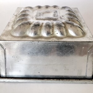May include: A vintage, rectangular, aluminum baking mold. The top features a decorative, raised floral design surrounding a square center. The sides are smooth, and the mold sits on a small, rectangular base. The metal has a slightly aged appearance.