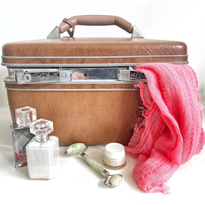 May include: A brown Samsonite suitcase with silver hardware, a brown handle, and a pink scarf draped over the side. Beside the suitcase are two perfume bottles, a jade roller, and a small jar of cream.