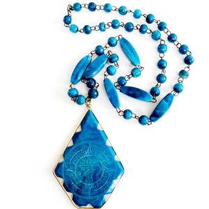 May include: A necklace featuring a large, diamond-shaped blue pendant with an embossed circular design, framed in gold-tone metal. The chain consists of alternating round and elongated blue beads connected by dark metal links.