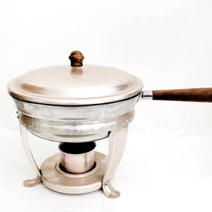May include: A vintage silver chafing dish with a lid and wooden handle. The dish sits on a metal stand with curved legs and a small burner compartment. The lid has a wooden knob.