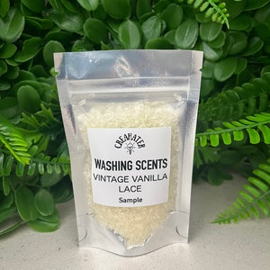 May include: A transparent, sealed plastic sachet containing off-white granules. The sachet has a white label with the text "WASHING SCENTS VINTAGE VANILLA LACE SAMPLE" and a logo. Green foliage forms the background.