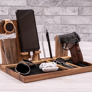 Può includere: Un organizzatore in legno con più scomparti per contenere vari oggetti, tra cui uno smartphone, un orologio, occhiali da sole, chiavi, auricolari e una penna. L'organizzatore è rivestito in feltro nero e ha una finitura in legno naturale.
