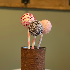 K&ouml;nnte beinhalten: Drei Cake Pops mit unterschiedlich gefrostetem Zuckerguss und Streuseln werden in einer rostigen Metalldose pr&auml;sentiert. Die Dose steht auf einem wei&szlig;en, mehrst&ouml;ckigen Kuchenst&auml;nder.