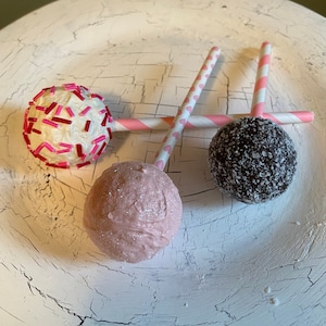 K&ouml;nnte beinhalten: Drei Cake Pops auf wei&szlig;en, gestreiften Stielen. Die Cake Pops sind mit rosa Streuseln, wei&szlig;em Frosting und Schokoladenstreuseln verziert.