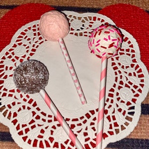 K&ouml;nnte beinhalten: Drei Cake Pops auf Stielen, verziert mit rosa Zuckerguss, roten und rosa Streuseln und Schokolade. Die Cake Pops befinden sich auf einer wei&szlig;en Serviette.