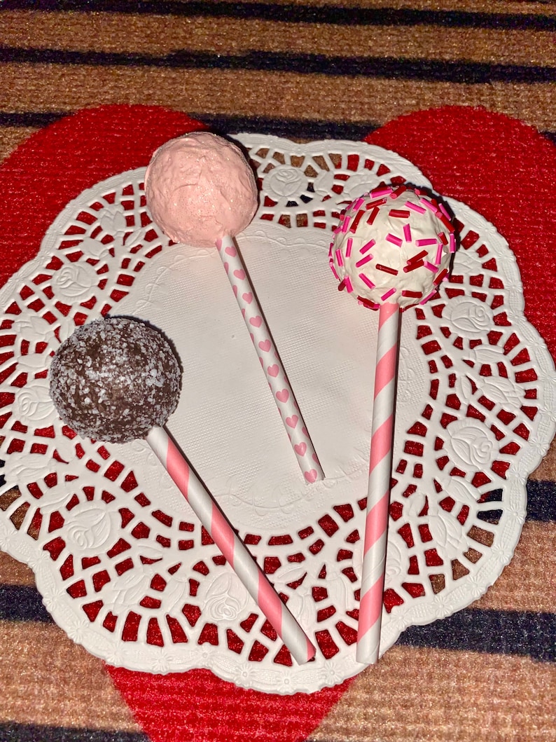 K&ouml;nnte beinhalten: Drei Cake Pops auf Stielen, verziert mit rosa Zuckerguss, roten und rosa Streuseln und Schokolade. Die Cake Pops befinden sich auf einer wei&szlig;en Serviette.