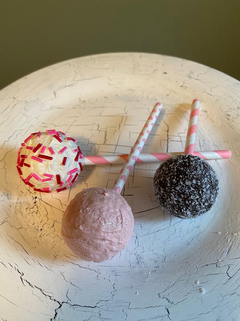 K&ouml;nnte beinhalten: Drei Cake Pops auf wei&szlig;en, gestreiften Stielen. Die Cake Pops sind mit rosa Streuseln, wei&szlig;em Frosting und Schokoladenstreuseln verziert.
