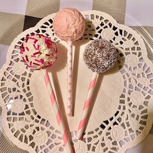 K&ouml;nnte beinhalten: Drei Cake Pops auf rosa-wei&szlig;en gestreiften Stielen. Die Cake Pops sind mit rosa, wei&szlig;em und braunem Frosting und Streuseln &uuml;berzogen. Die Cake Pops befinden sich auf einer wei&szlig;en Serviette.
