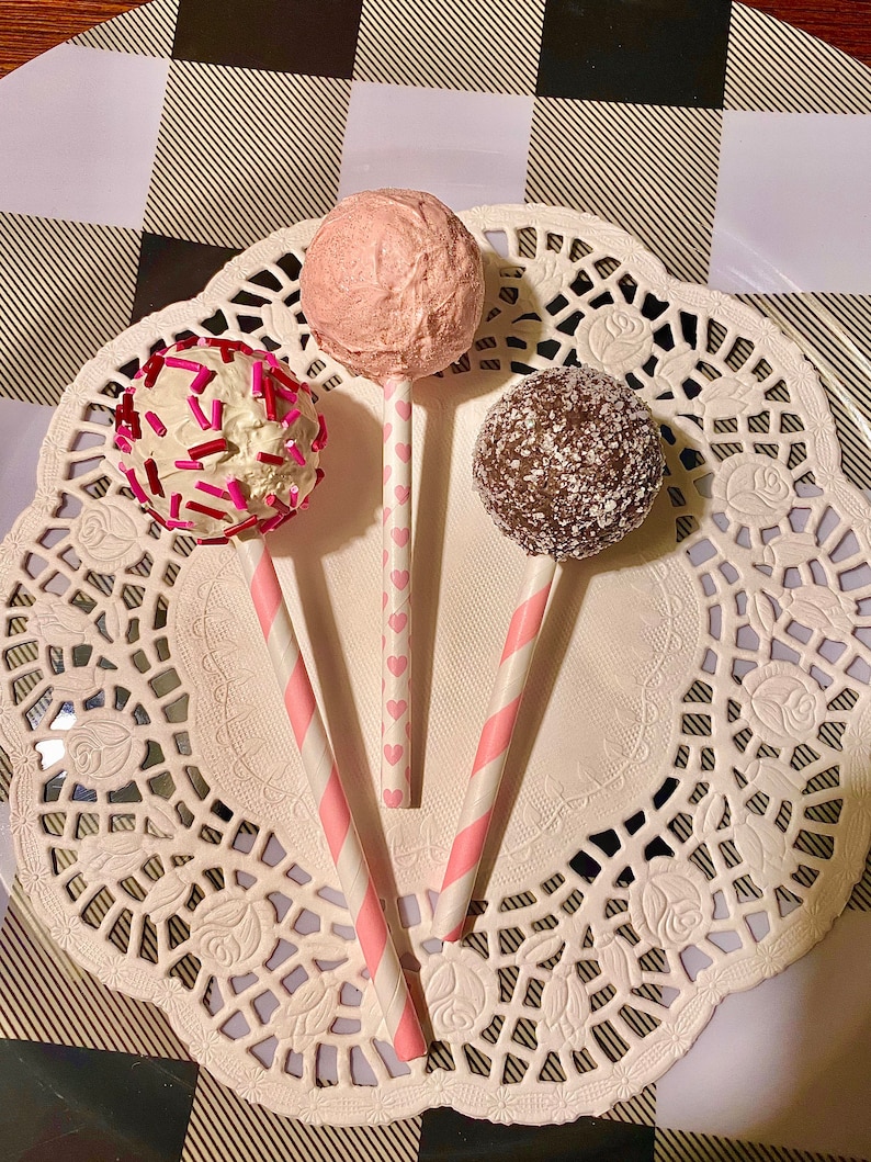 K&ouml;nnte beinhalten: Drei Cake Pops auf rosa-wei&szlig;en gestreiften Stielen. Die Cake Pops sind mit rosa, wei&szlig;em und braunem Frosting und Streuseln &uuml;berzogen. Die Cake Pops befinden sich auf einer wei&szlig;en Serviette.