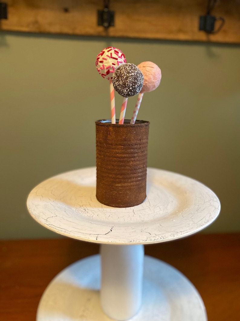 K&ouml;nnte beinhalten: Drei Cake Pops mit unterschiedlich gefrostetem Zuckerguss und Streuseln werden in einer rostigen Metalldose pr&auml;sentiert. Die Dose steht auf einem wei&szlig;en, mehrst&ouml;ckigen Kuchenst&auml;nder.