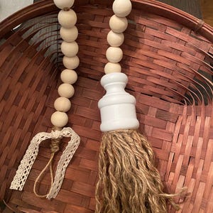 May include: A decorative wooden bead garland with a white painted accent piece and a jute tassel. The garland also features a lace ribbon. The beads are light brown and the tassel is a natural brown color.