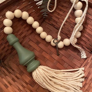 May include: Decorative wooden bead garland with a green painted finial and a cream-colored tassel. The garland includes a small, antique-style key and is displayed on a woven basket.