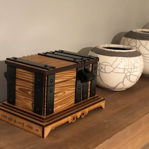 May include: A wooden treasure chest with black metal accents and a decorative base. The chest features a light brown wood grain pattern and is adorned with black metal hinges and handles. The base has carved details and is displayed on a wooden shelf.