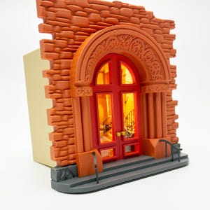 May include: A miniature brick building with a red double door and a decorative archway. The door has glass panels and a small staircase behind it. The building is made of orange brick and has a gray base.