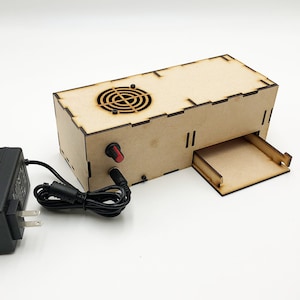 Op de afbeelding: Een houten doos met een klein lade en een ronde ventilator erin. De doos is verbonden met een zwarte adapter met een snoer.
