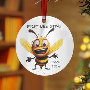 May include: A white, round ornament with a cartoon bee illustration. The bee is yellow and black with outstretched wings and a smiling face. Text on the ornament reads "FIRST BEE STING" and "SAM 2024". A red ribbon is attached for hanging.