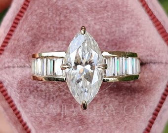 May include: A gold ring featuring a marquise-cut diamond and baguette-cut side stones. The ring is presented in a pink velvet box, with a backdrop of green foliage.
