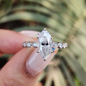 Könnte beinhalten: Ein Verlobungsring mit Diamanten im Marquiseschliff und einem Weißgoldband. Der Ring hat einen großen, zentralen Diamanten und kleinere Akzentsteine. Das Band hat ein zartes Design mit abwechselnd runden und marquiseförmigen Steinen. Der Ring wird vor einem verschwommenen grünen Hintergrund gehalten.