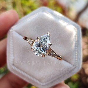 May include: A gold engagement ring with a pear-shaped diamond set in a velvet ring box. The ring features smaller accent diamonds on the band. The ring is held in a hand, with a blurred natural background.