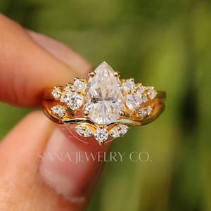 May include: A gold ring featuring a central pear-shaped clear gemstone surrounded by smaller round clear stones in a cluster arrangement. The band is polished gold, and the text SANA JEWELRY CO. is visible on the surface of the ring setting.