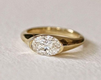 May include: A gold ring with a large, oval-cut diamond. The ring has a smooth, polished band and a bezel setting that securely holds the diamond. The diamond sparkles brightly, reflecting light. The ring is a piece of jewelry.