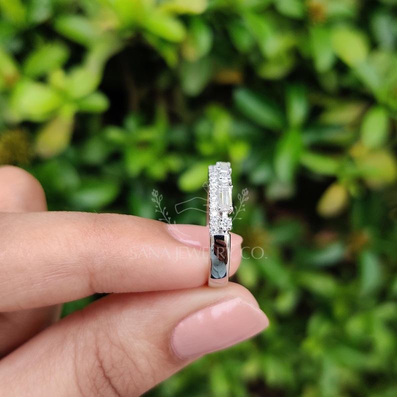 May include: A silver ring with a row of small, round diamonds and a row of baguette-cut diamonds. The ring is held in a person's hand, with a blurred green background.