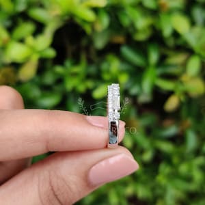 May include: A silver ring with a row of small, round diamonds and a row of baguette-cut diamonds. The ring is held in front of a green, leafy background. The ring is a piece of jewelry.