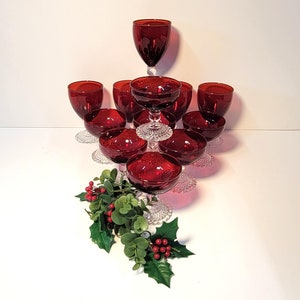May include: A collection of ruby red glassware, including wine glasses and stemmed bowls, arranged on a white surface. The glasses have a classic design. Green and red holly and berries are arranged at the base of the glasses.