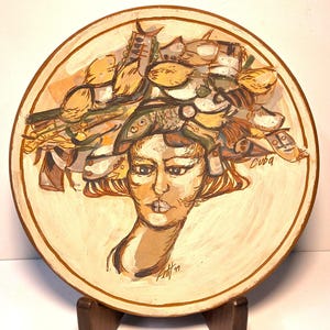 May include: A decorative ceramic plate featuring a painted portrait of a woman with an elaborate headdress. The artwork is in shades of beige, brown, and yellow, with the word "Cuba" visible. The plate is displayed on a wooden stand.