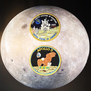 May include: Two embroidered patches featuring space-themed designs. The top patch commemorates the "First Lunar Landing of Mankind 1969" with a lunar module illustration. The bottom patch displays an eagle and the words "Apollo 11". Both patches are set against a backdrop resembling the moon.