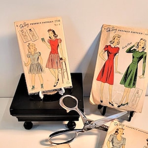 May include: Four vintage sewing patterns featuring women's dresses. The patterns are from the 1940s and 1950s and are in good condition. The patterns are all different styles and include a red dress, a green dress, a blue dress, and a plaid dress. The patterns are all on white paper with black and white illustrations.