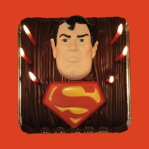 May include: A chocolate cake with a Superman logo and a sculpted Superman head on top. The cake is decorated with red candles.