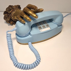 May include: A light blue vintage telephone with a gold-colored trim and a coiled handset cord. A gold-colored hand sculpture rests on the phone. The phone has a keypad with numbers and symbols.