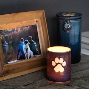 May include: A wooden picture frame containing a photo of two people and a dog sitting on a bench by a lake. A ceramic candle holder with a paw print design is lit and sitting on a wooden surface next to a blue ceramic urn with a paw print lid.