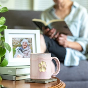 May include: A pink ceramic mug with a gold paw print design. The mug is sitting on a grey coaster on a wooden table. A framed photo of a woman and a dog is in the background.
