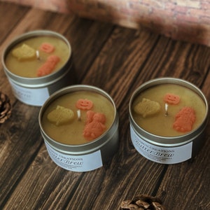 May include: Three small, silver tin candles with a pale yellow wax. Each candle features decorative wax shapes of a pumpkin, leaf, and gingerbread man. The label on the candle reads "Butter Brew".