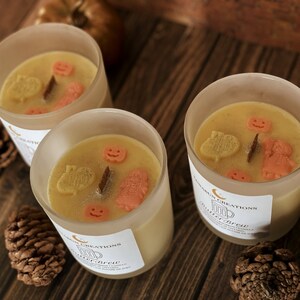May include: Three glass candles with a light yellow wax, decorated with orange and yellow shapes, including pumpkins and leaves. Each candle has a label with the text "Butterbrew". The candles are on a wooden surface with pinecones and pumpkins.