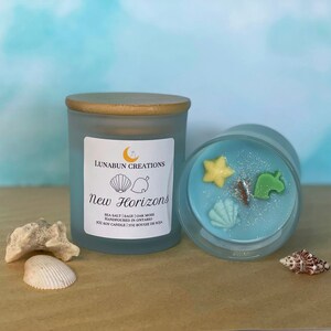 Pode incluir: Uma vela de soja azul em um frasco de vidro fosco com tampa de madeira. A vela é decorada com uma concha branca, uma folha verde e o texto "New Horizons". A vela é cercada por conchas e um fundo azul claro.