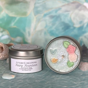 Op de afbeelding: Twee zilverkleurige blikken kaarsen. Eén kaars heeft een label met de tekst "Fairy Fountain" en "Lunabun Creations". De andere kaars heeft een decoratieve bovenkant met bloemen en een vlinder. Het nettogewicht is 113 gram.