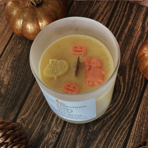 May include: A candle in a clear glass jar with a light yellow wax, decorated with small orange pumpkin and figure-shaped wax pieces. The candle sits on a wooden surface with gold pumpkins and pinecones. The label reads "Autumn Creations".